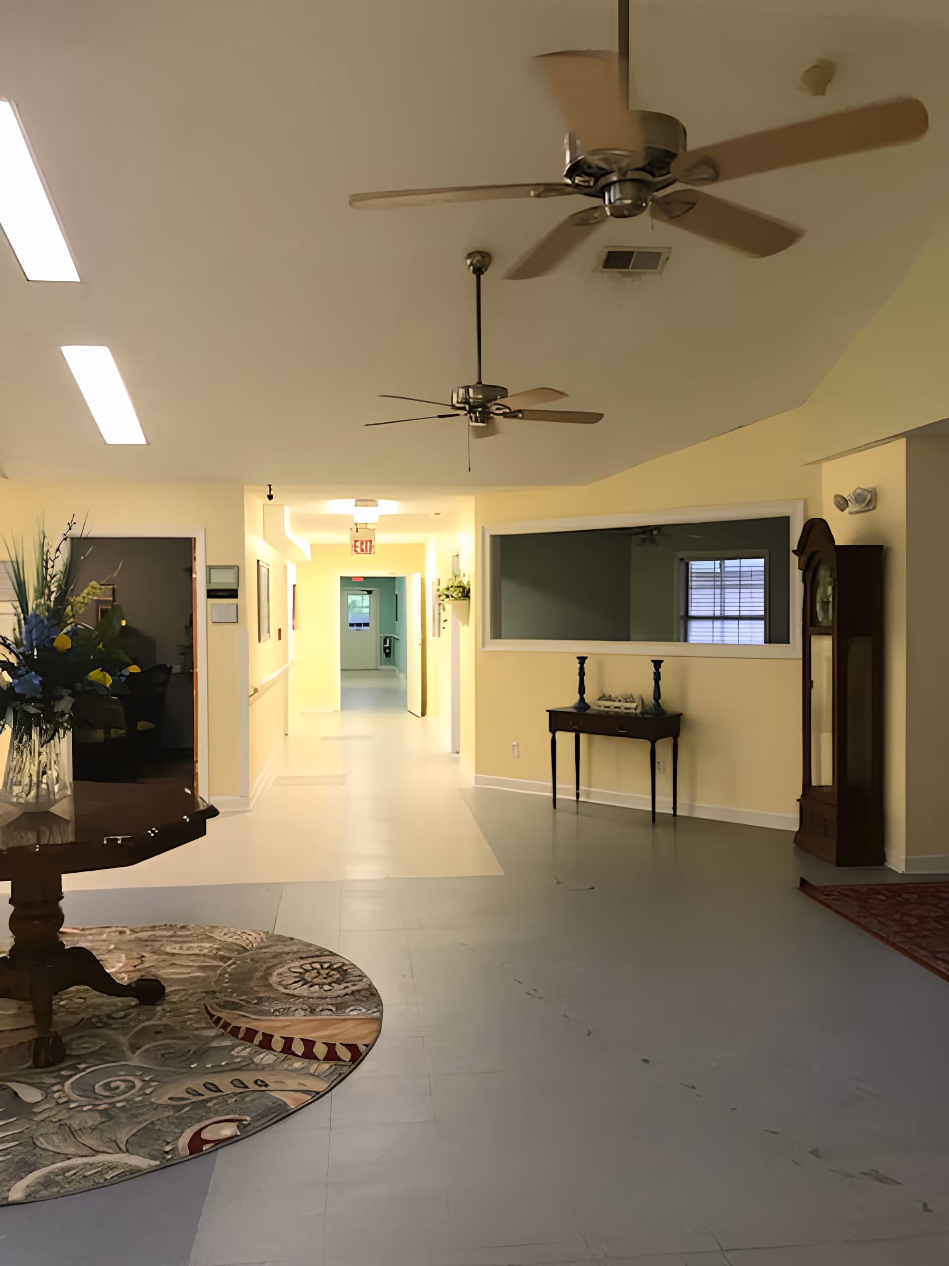 Interior lobby/hallway of an assisted living facility with ceiling fans, a table with flowers, a small side table and a grandfather clock leading to a corridor and exit sign.