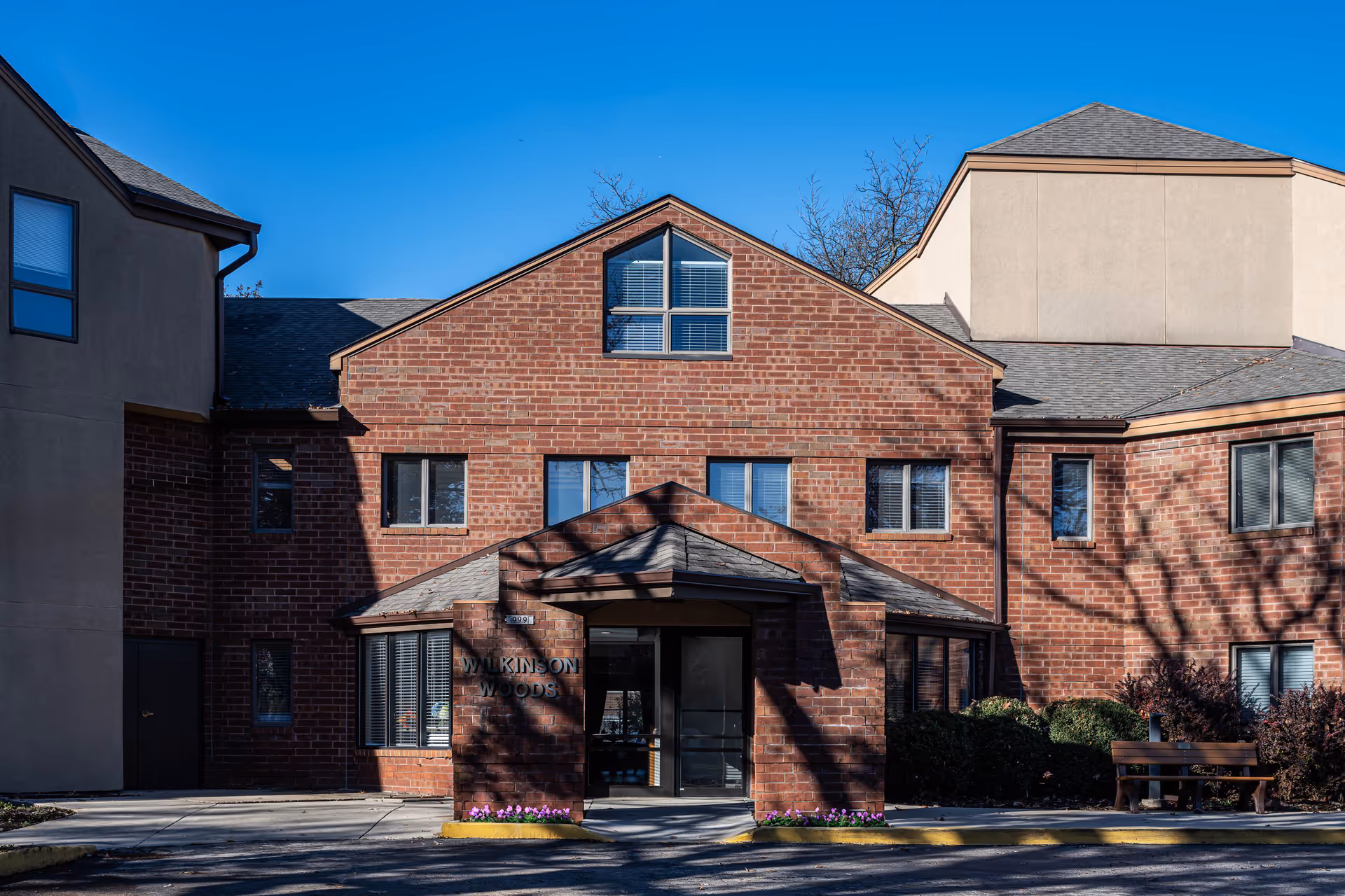 Front exterior view of Wilkinson Woods Senior Community building with brick facade, multiple windows, and a small covered entrance. There is a bench and some bushes on the right side, and the sky is clear and blue.
