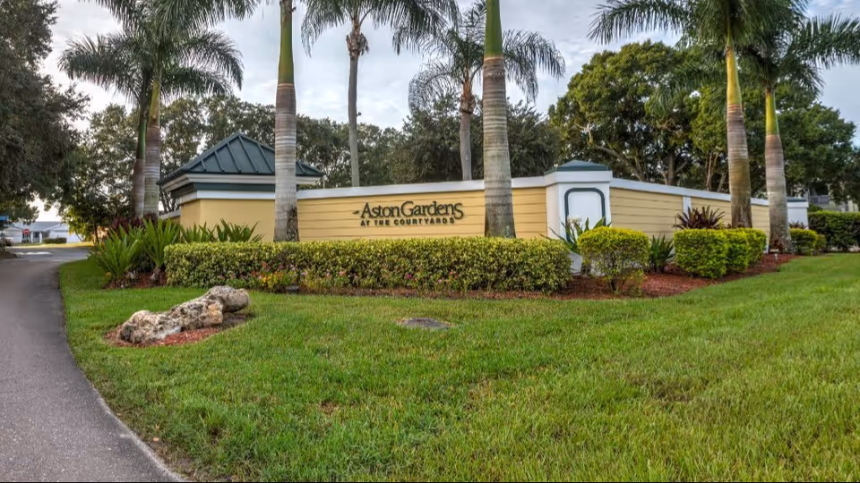 Entrance sign for Aston Gardens at the Courtyards surrounded by palm trees, shrubs, and a manicured lawn.
