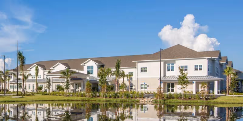 Two-story senior living facility building with palm trees and landscaping reflected in a pond under a blue sky.