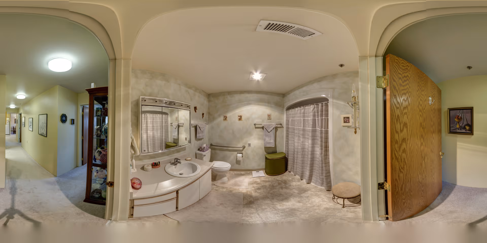 Wide panoramic interior showing a bathroom with a vanity and sink, toilet, shower with curtain, and an adjacent hallway with an open wooden door.