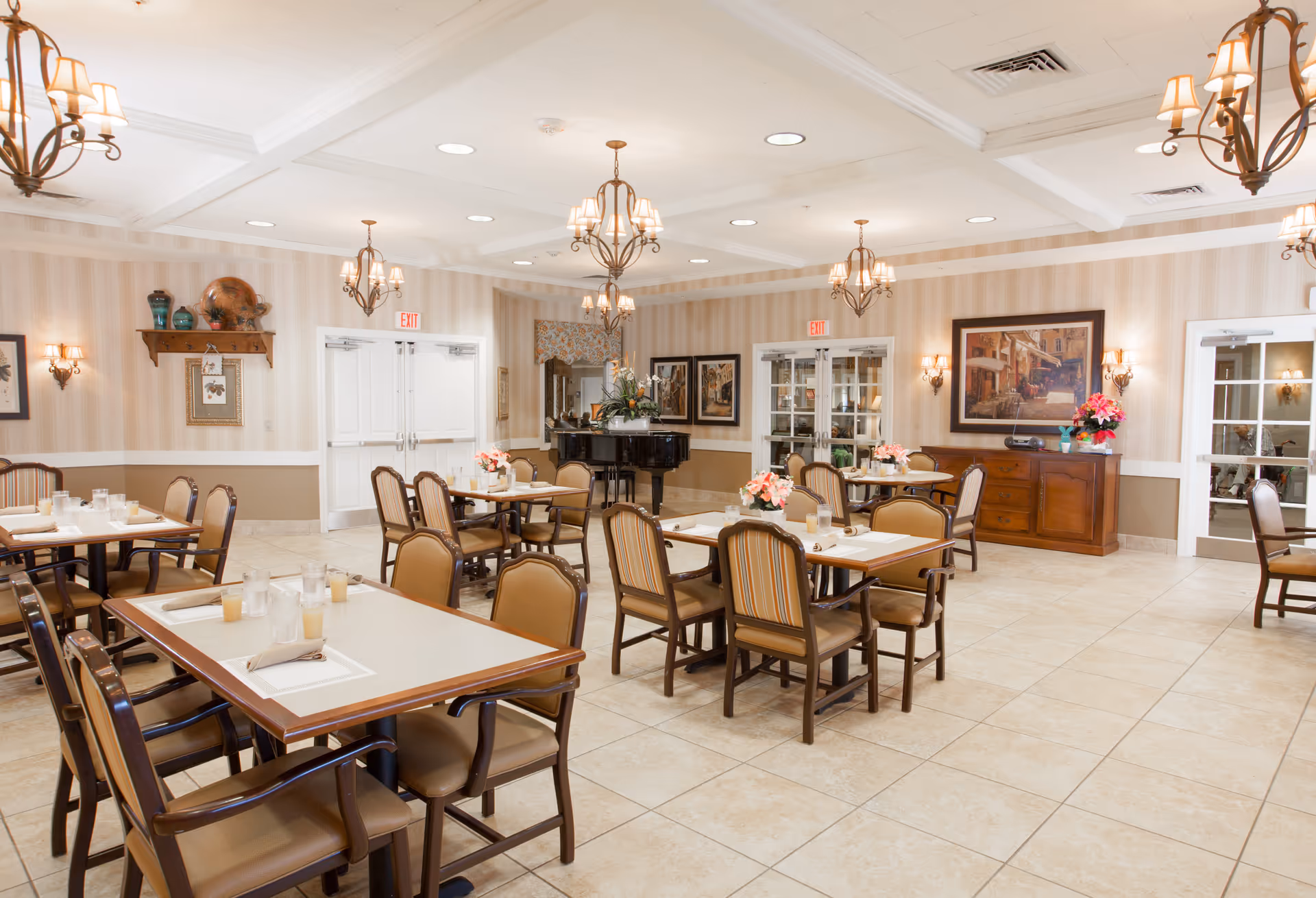 Spacious, well-lit dining room with several set tables and chairs, chandeliers, artwork, and a piano.