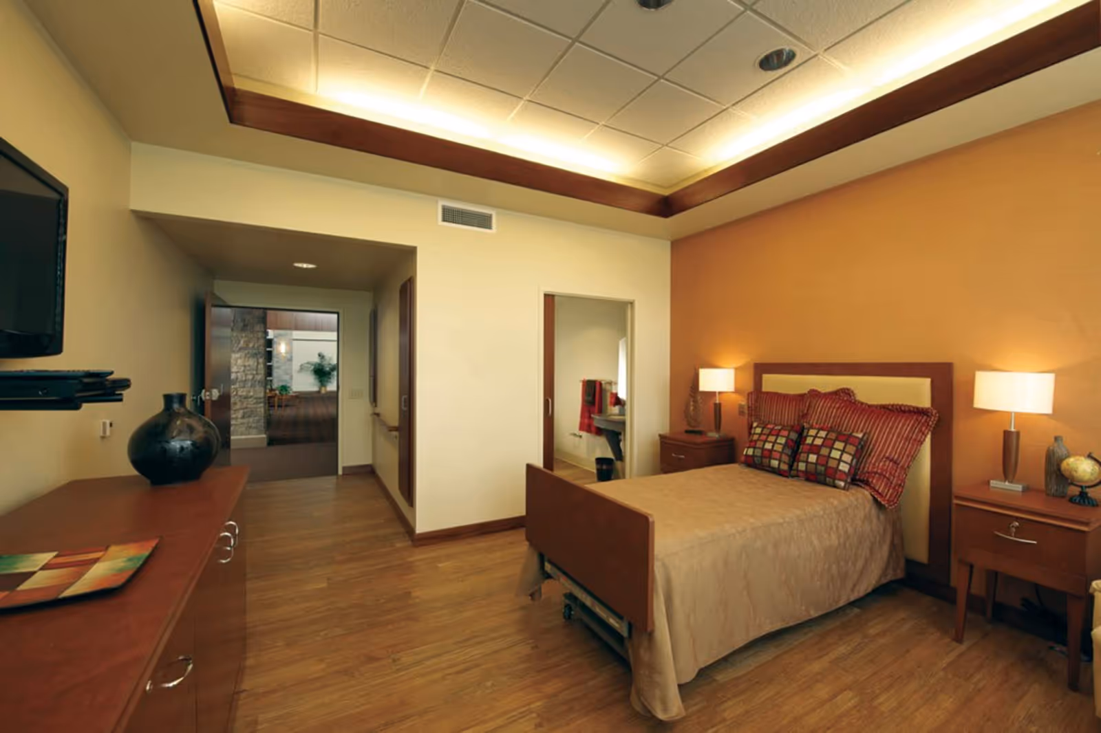 A cozy bedroom in a senior living facility with a single bed covered in a beige quilt and decorative pillows. The room features wooden flooring, two bedside tables with lamps, a dresser with a decorative vase, and a wall-mounted TV. The walls are painted in warm tones, and there is an open doorway leading to another area.