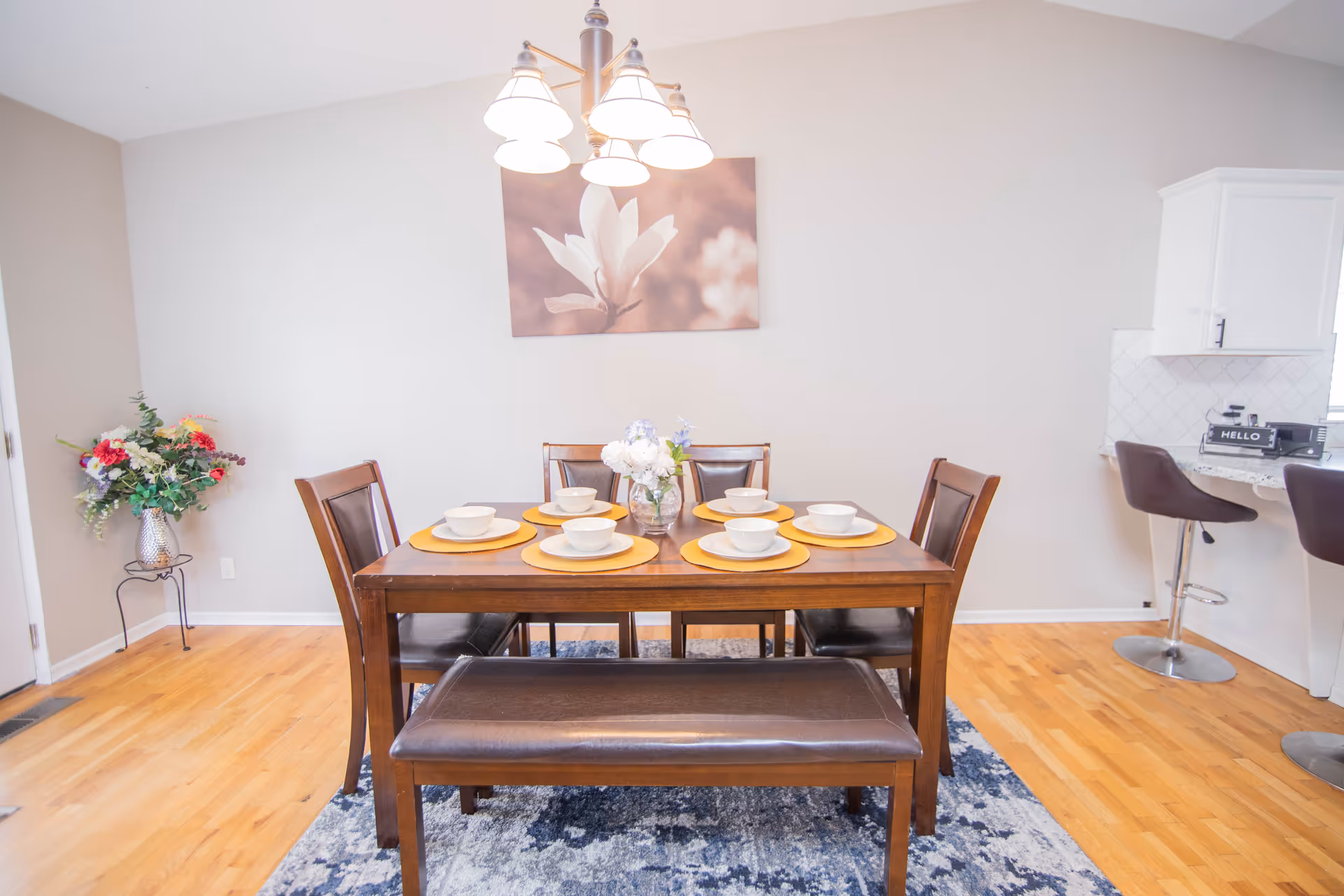 A dining area with a wooden table set for six, including four chairs and a bench. The table has yellow placemats, white bowls, and a vase with flowers in the center. Above the table hangs a chandelier with five lights. There is a floral painting on the wall behind the table, a flower arrangement in a vase on a small stand to the left, and two bar stools at a kitchen counter to the right.