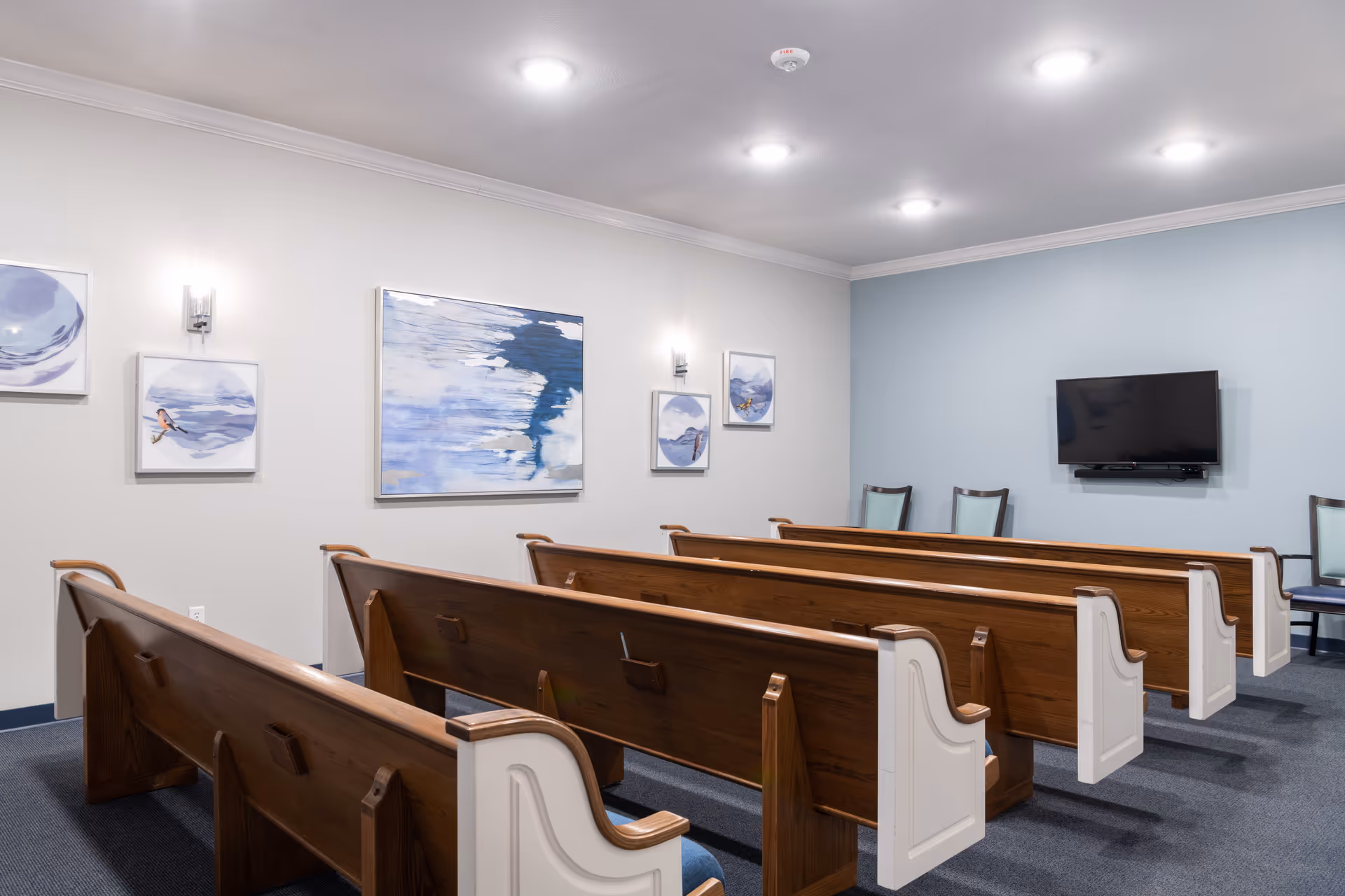 Empty chapel-style room with wooden pews facing a wall-mounted TV and framed abstract artwork.