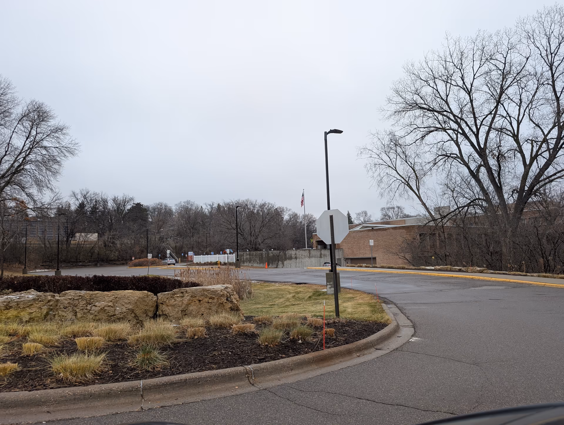 Outdoor view of a parking lot area with a curved road, some large rocks, leafless trees, and a building in the background under an overcast sky. An American flag is visible near the building.