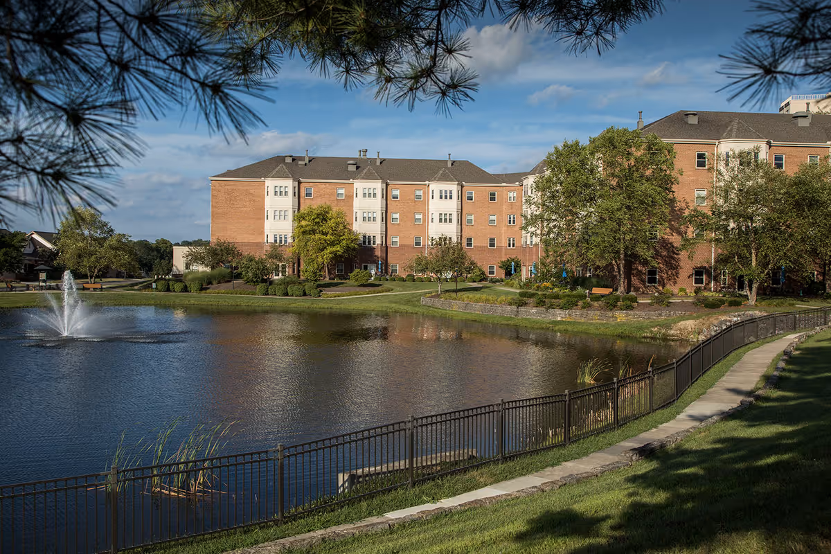 A large brick senior living facility building named Bethany Village is seen behind a pond with a water fountain. The pond is surrounded by a black metal fence and a paved walkway. Trees and green grass are visible around the building and pond under a partly cloudy blue sky.