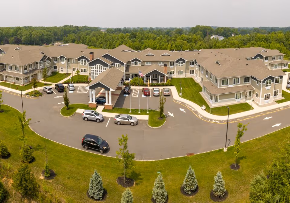 Aerial view of Juniper Village at Hamilton, showing a large senior living facility building with multiple wings, a covered entrance, several parked cars, and surrounding green lawns and trees.