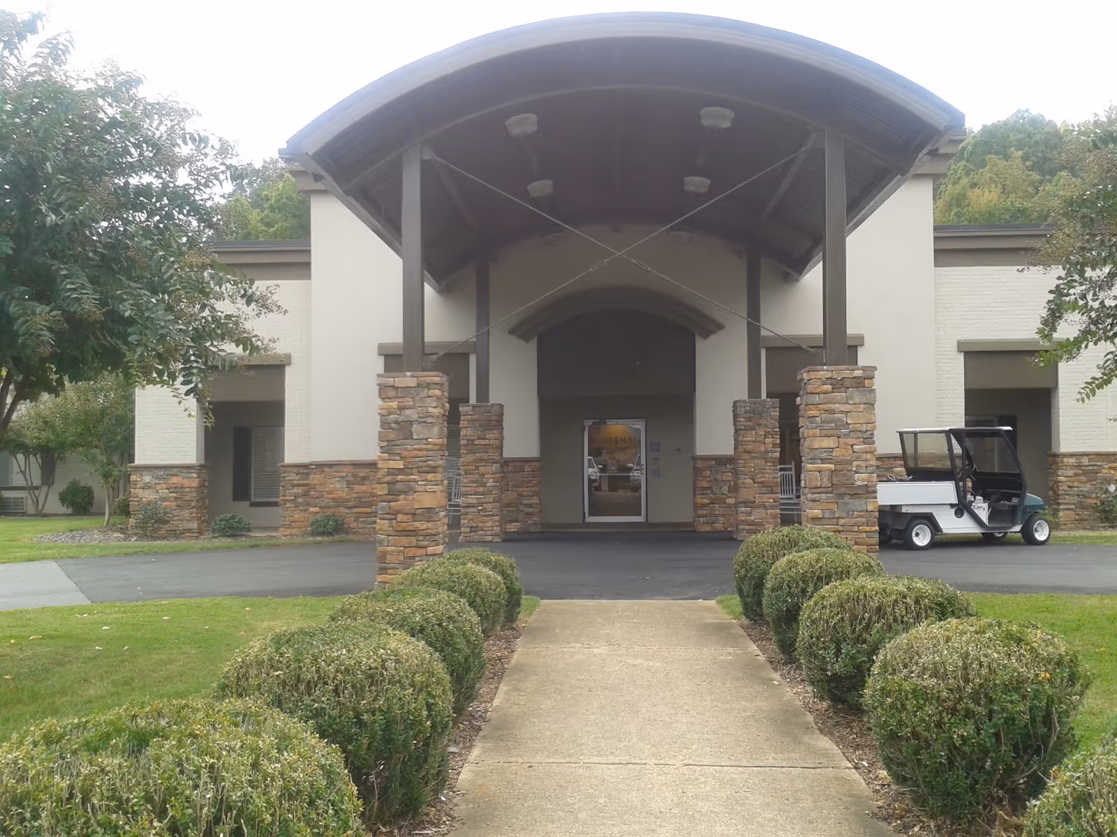 Entrance of a building with a covered driveway supported by stone pillars, a concrete walkway lined with trimmed bushes leading to the door, and a small white utility vehicle parked on the right side. Trees and grass surround the area.