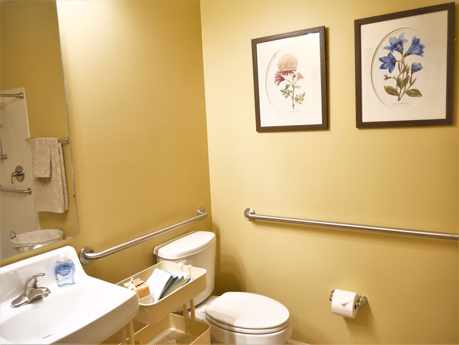 A bathroom with yellow walls featuring a white toilet and sink. There are two framed floral pictures on the wall above a horizontal grab bar. A towel rack with a beige towel is visible in the mirror reflection. A small cart next to the toilet holds folded towels and soap. A bottle of Softsoap hand soap is on the sink.