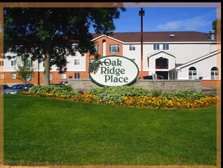 Exterior view of Oak Ridge Place facility with a large sign displaying the name in front of a well-maintained lawn and flower bed, with the building visible in the background under a clear blue sky.