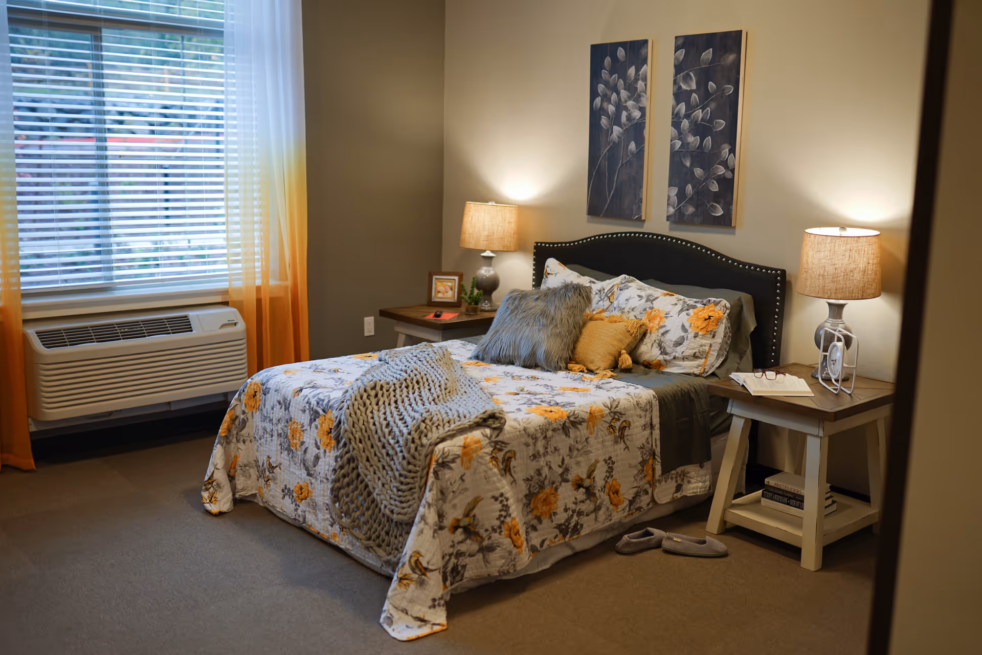 A cozy bedroom with a floral-patterned bed, nightstands and lamps, window with blinds and an air conditioning unit.