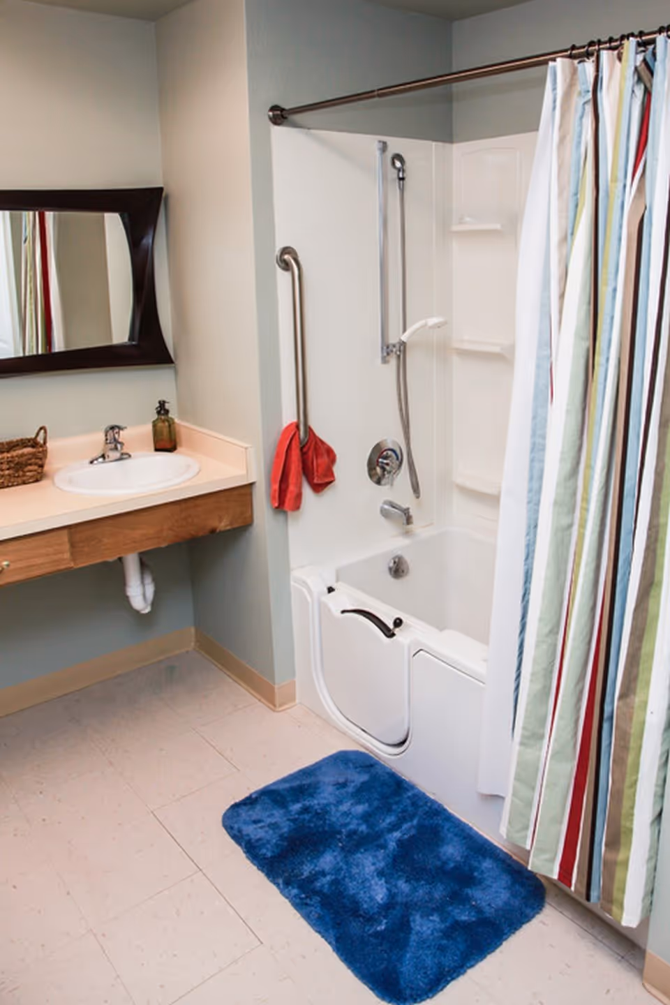Bathroom with a walk-in bathtub featuring a safety handle and a handheld showerhead. There is a striped shower curtain, a blue bath mat on the floor, a sink with a mirror above it, and a red towel hanging on a grab bar next to the tub.