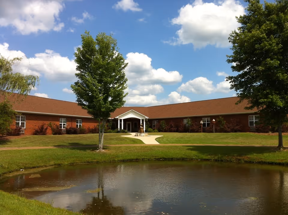A single-story brick building with a brown roof, surrounded by green grass and trees, under a partly cloudy blue sky. In front of the building is a small pond reflecting the trees and sky. A paved walkway leads to the building's entrance, which has a white portico.