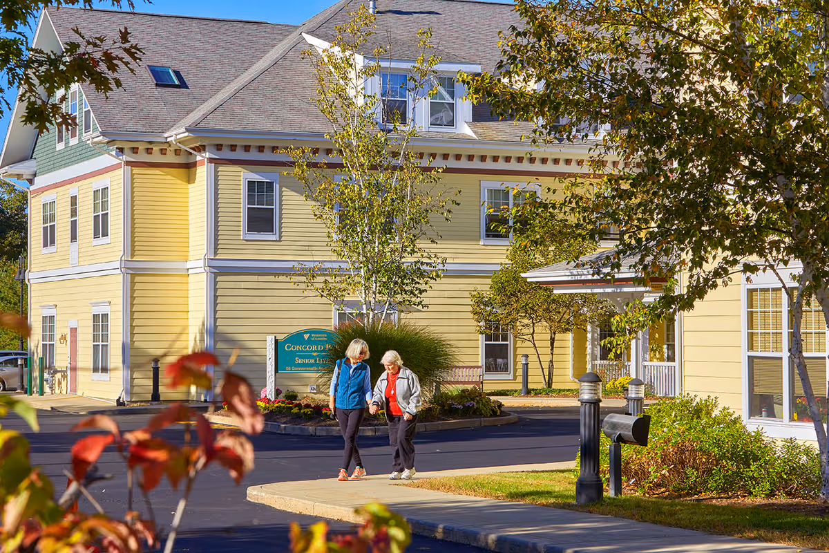 Two people walking near the entrance of a yellow, multi-story senior living building surrounded by landscaping.