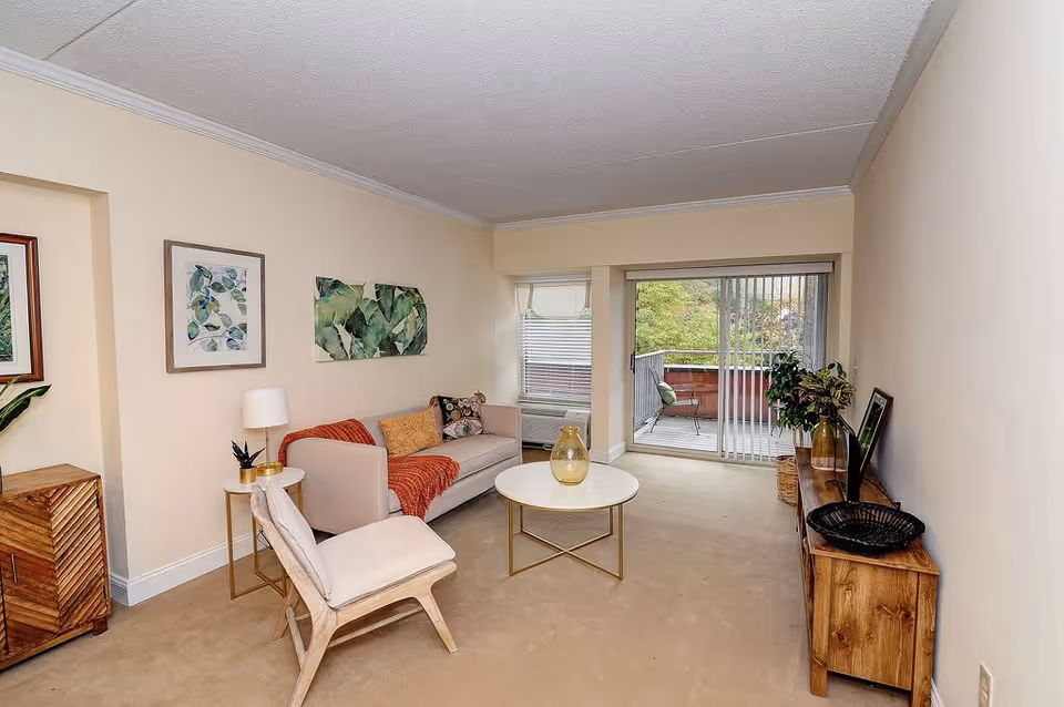 A cozy living room with beige walls and carpet, featuring a light beige sofa with colorful cushions and a red throw blanket, a wooden chair with a white cushion, a round white coffee table with a glass vase, a wooden sideboard with decorative items, and framed botanical artwork on the walls. Sliding glass doors lead to a small balcony with outdoor seating and greenery visible outside.