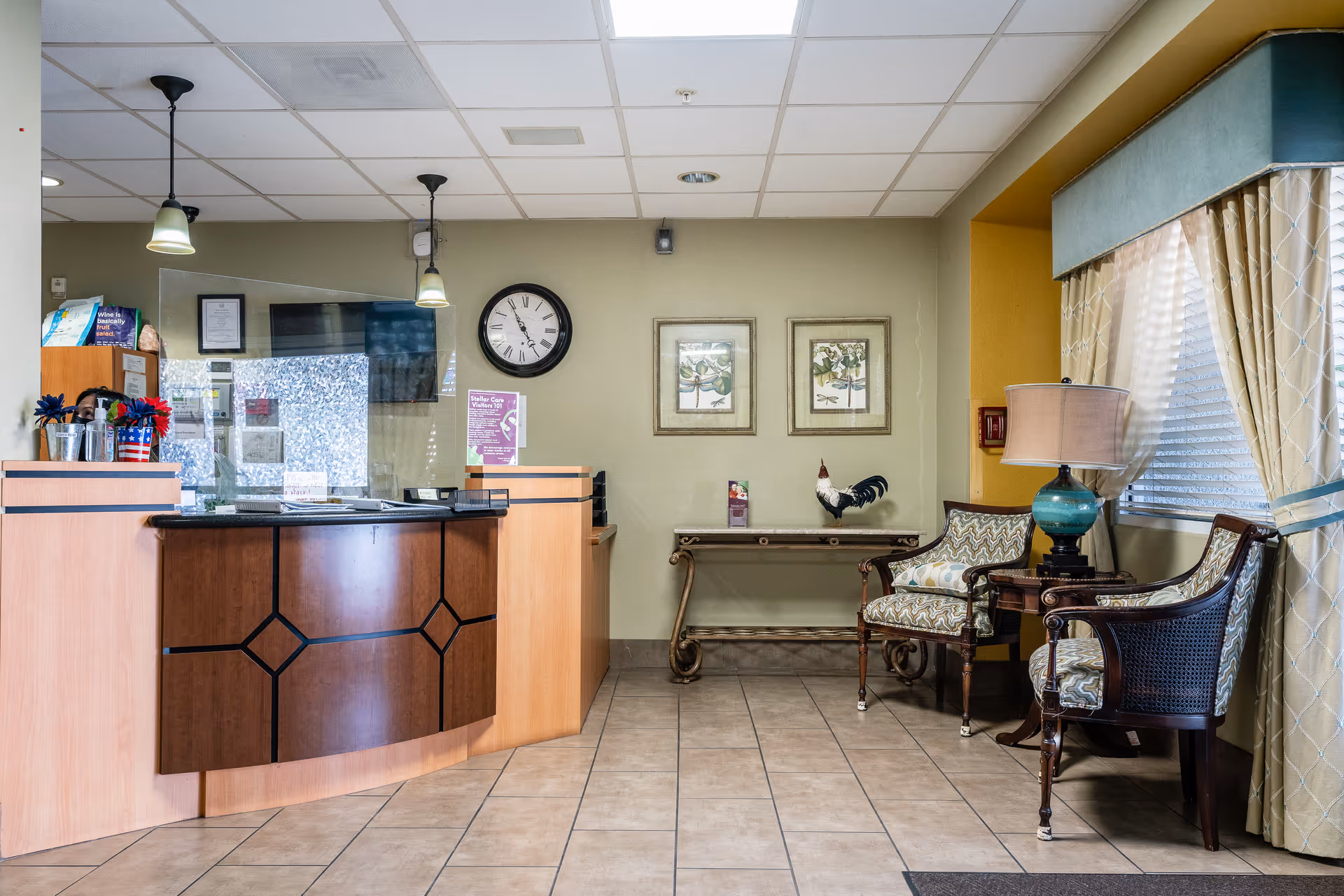 Reception area of a senior living facility showing a curved front desk, wall clock, decorative table, and two upholstered chairs by a window.