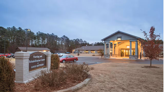 Exterior view of the Nursing and Rehabilitation Center at Good Shepherd during dusk, showing the building entrance with a covered drop-off area, a parking lot with several cars, a sign with the facility name, and trees in the background.