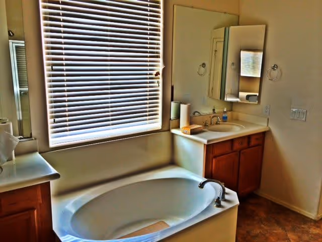 A bathroom featuring a white bathtub with a silver faucet beneath a window with closed blinds. To the right of the bathtub is a countertop with a sink, a mirror above it, and wooden cabinets below. The walls are light-colored and there is a towel ring mounted on the wall near the sink.