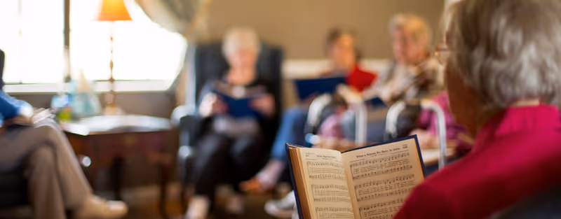 A group of elderly people sitting in a living room, with one person in the foreground holding and reading from a music book. The others are seated in chairs, some holding songbooks, in a cozy, well-lit room with a lamp and window in the background.