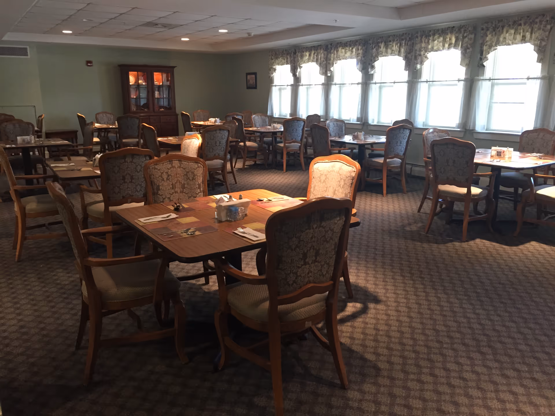 A dining room with multiple wooden tables and upholstered chairs arranged neatly. The tables are set with placemats, napkins, and cutlery. Large windows with floral valances allow natural light to fill the room. A wooden cabinet with glass doors is visible against the far wall.