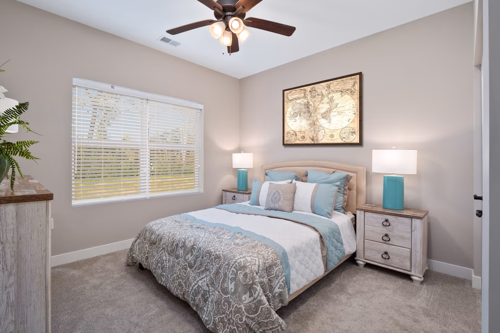 A neatly made bed with patterned bedding and multiple pillows in a bedroom with beige walls, a ceiling fan with lights, two bedside tables with turquoise lamps, a framed vintage world map on the wall, a window with white blinds, and a dresser with a plant on top.