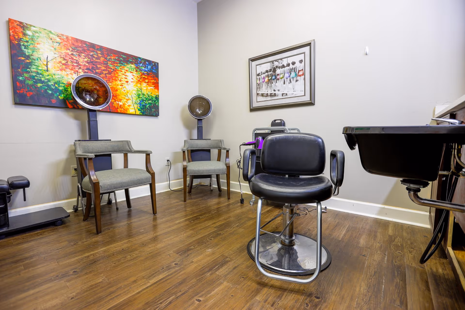 Interior view of a salon area with a black salon chair in the center, two gray chairs with hair dryers behind them, a black sink on the right, and colorful artwork on the wall.