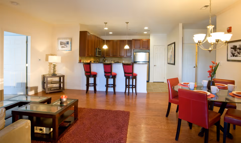 A bright and spacious open-concept living area featuring a kitchen with dark wood cabinets, stainless steel appliances, and a breakfast bar with three red cushioned stools. Adjacent to the kitchen is a dining area with a round glass table set with red chairs, plates, and cups. The living room area includes a beige sofa, glass-top coffee tables, a red rug, and a side table with a lamp. The walls are decorated with framed pictures, and a chandelier hangs above the dining table.