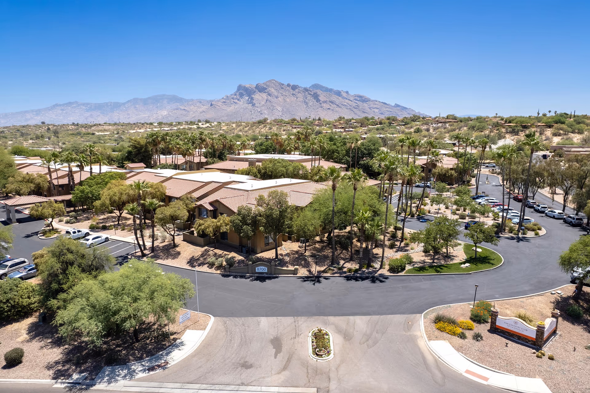 Aerial view of the Country Club of La Cholla senior living complex with low-rise buildings, palm trees, parking areas and mountains in the background.