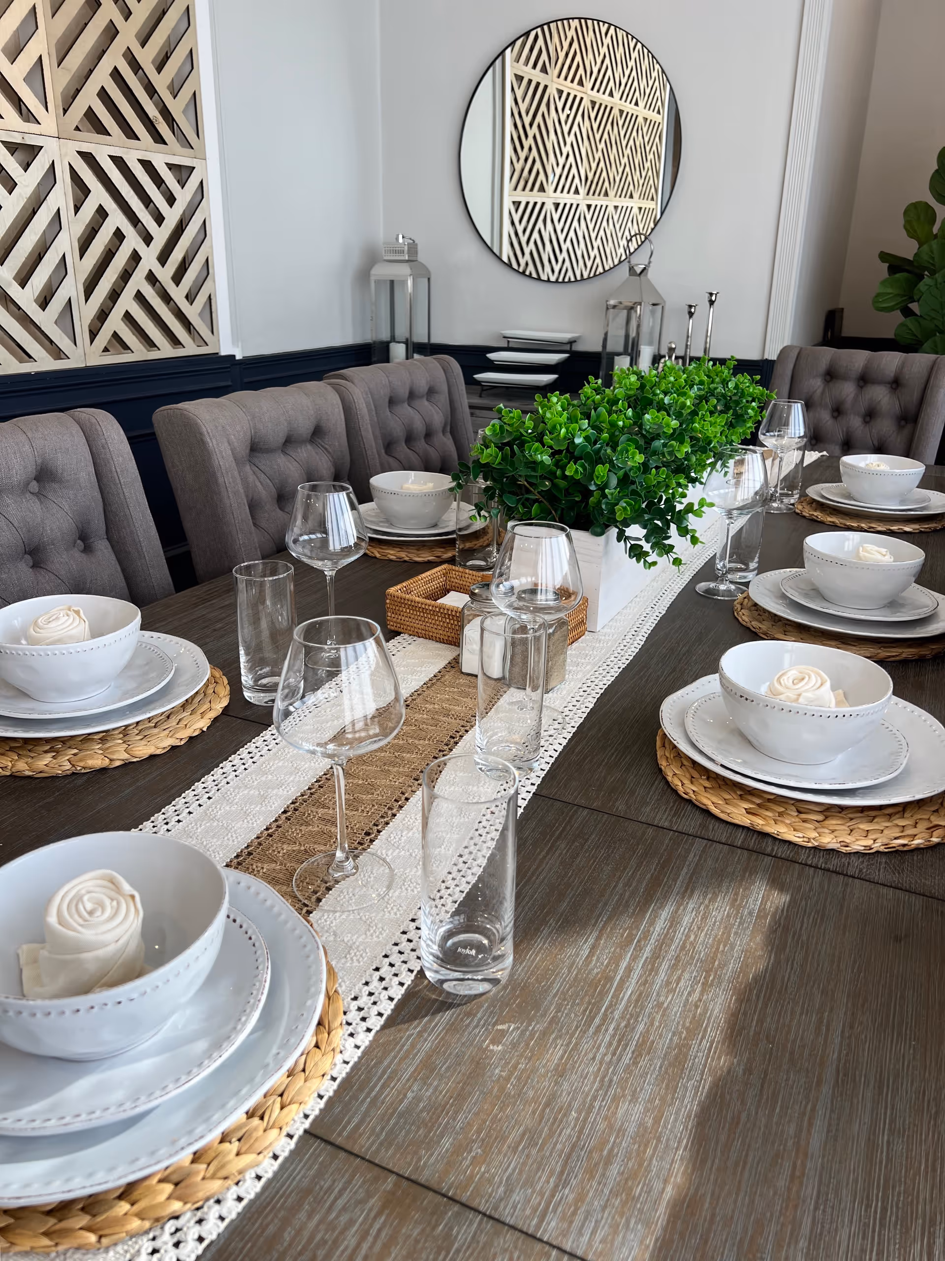A dining table set for six with woven placemats, white plates, bowls with rolled napkins, clear drinking glasses, and wine glasses. A long white planter with green plants serves as a centerpiece. The background features tufted gray chairs, a round wall mirror, decorative lanterns, and a geometric wooden wall panel.