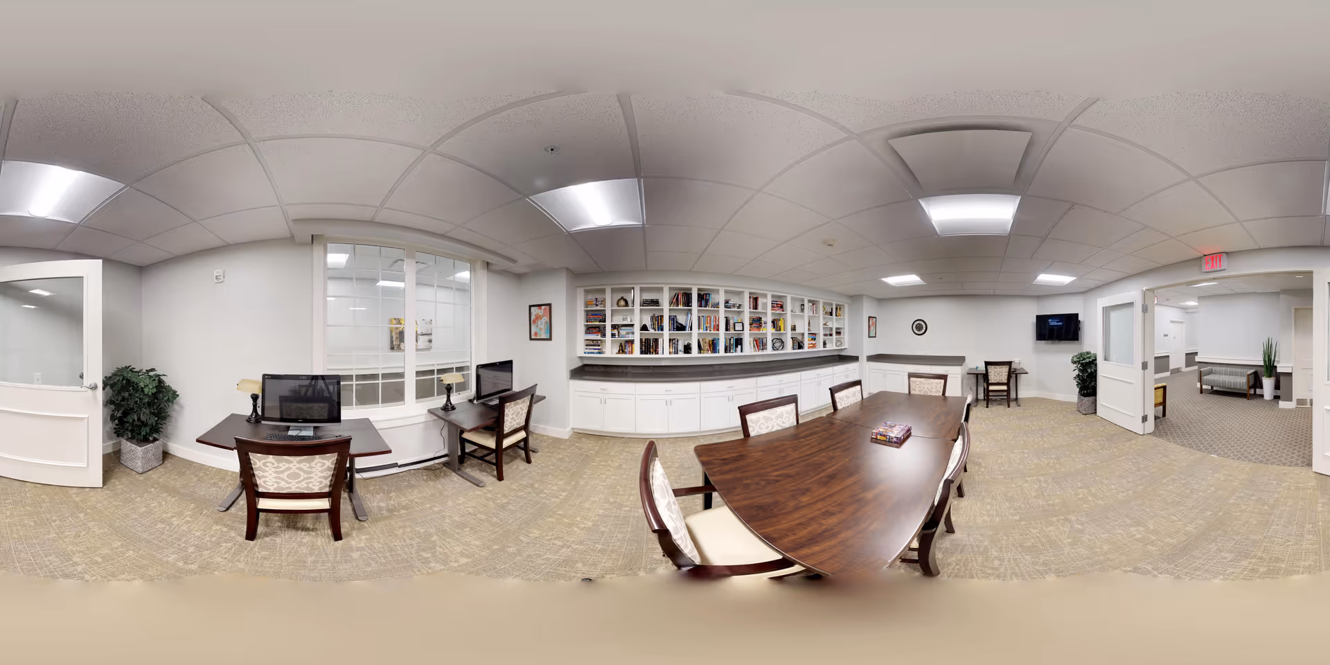 A spacious, well-lit common area in a senior living facility featuring a long wooden table with chairs around it, two desks with computers and chairs near a window, built-in white cabinets with shelves filled with books and decorative items, and an adjacent room visible through open double doors. The room has beige carpet, white walls, and ceiling lights.
