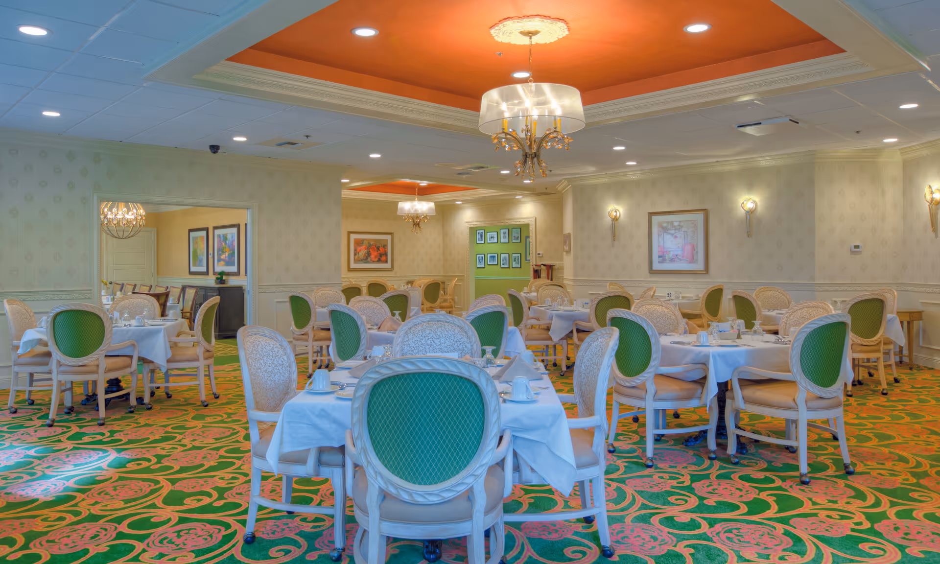 A brightly lit dining room with multiple round tables covered with white tablecloths, each set with cups, glasses, and napkins. The chairs have green cushioned backs and beige seats. The room features a colorful green and pink patterned carpet, cream-colored walls with framed artwork, and orange recessed ceiling sections with chandeliers.