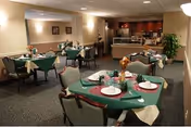 A dining room with multiple tables covered in green tablecloths and set with plates, glasses, and napkins. The room has beige walls, carpeted floors, and soft lighting. In the background, there is a counter area with cabinets and a plant near the wall.