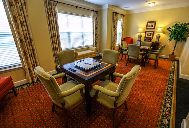A cozy common area with two sets of tables and chairs. The foreground features a square table with four cushioned chairs around it and a board game on top. The background has another table with four chairs, two table lamps, framed pictures on the wall, and a potted plant. The room has large windows with floral curtains and a red patterned carpet.