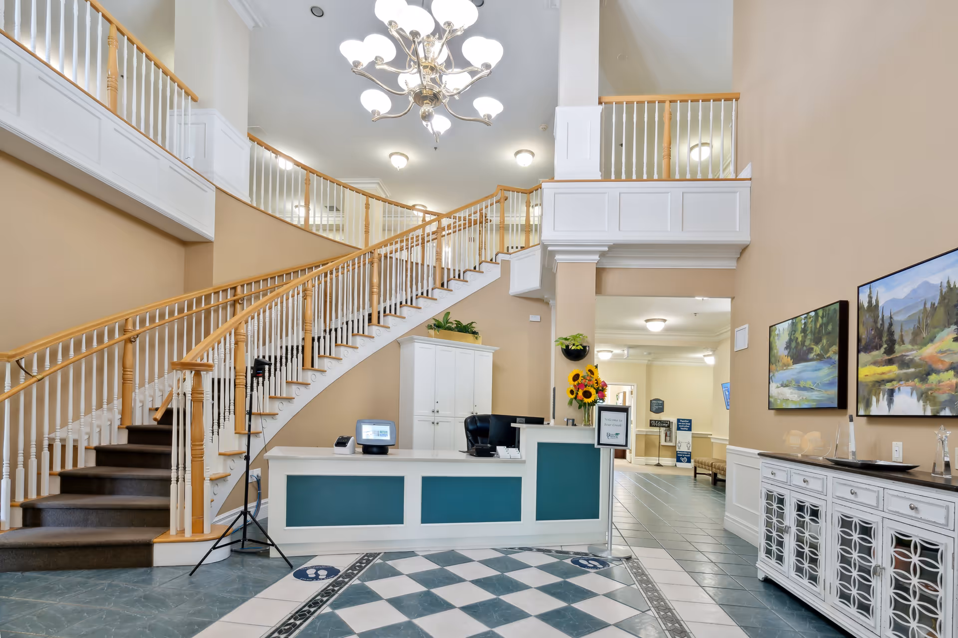 Interior view of Bear Creek Assisted Living lobby featuring a reception desk with computer and flowers, a curved staircase with wooden handrails and white balusters, a chandelier hanging from the ceiling, and framed landscape paintings on the wall.