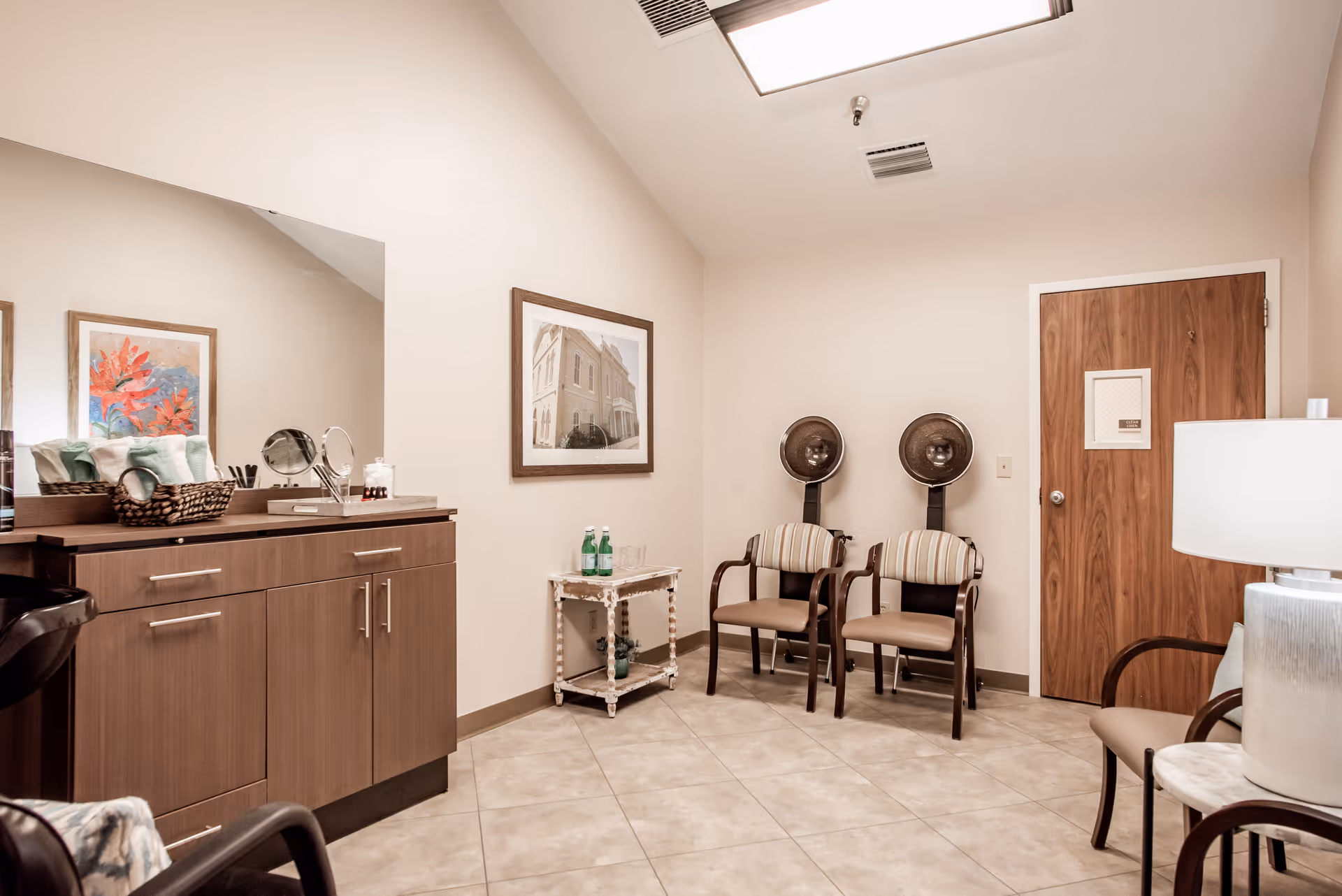 Interior view of a salon or beauty treatment room with two vintage hair dryers on stands, two striped chairs, a wooden cabinet with a large mirror, a small table with bottled water, and a wooden door. The room has tiled floors and neutral-colored walls with framed artwork.