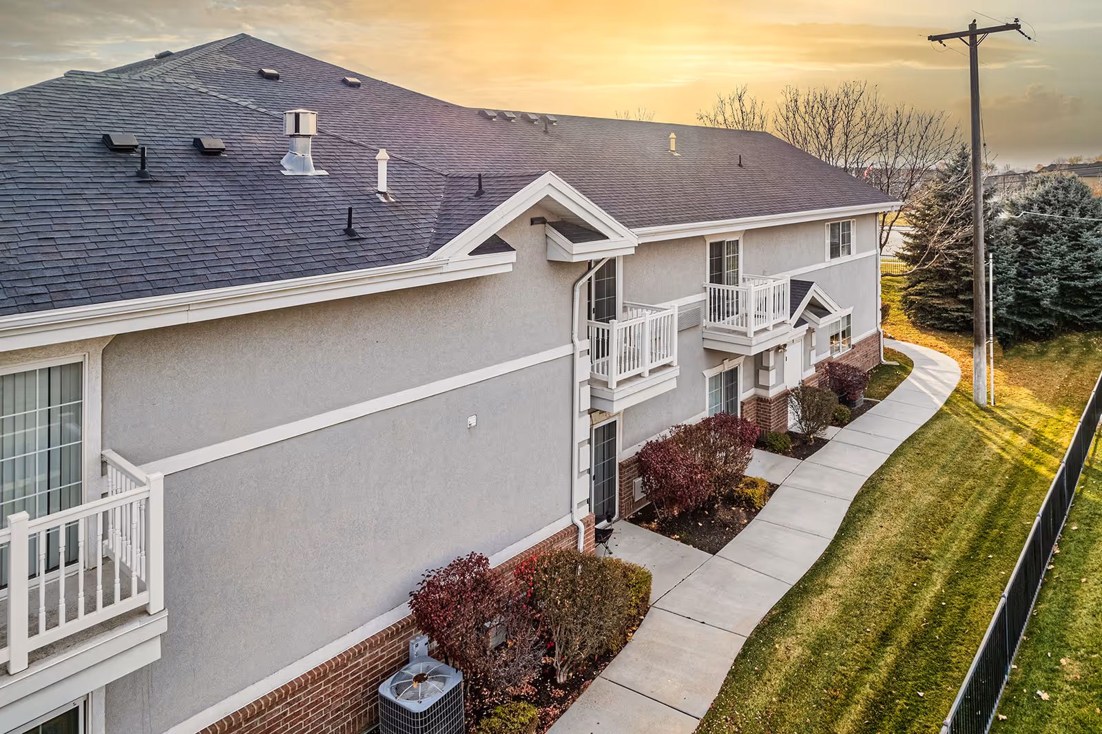 Exterior view of a two-story apartment-style senior living building with small balconies, a curved sidewalk, and landscaped lawn at sunset.
