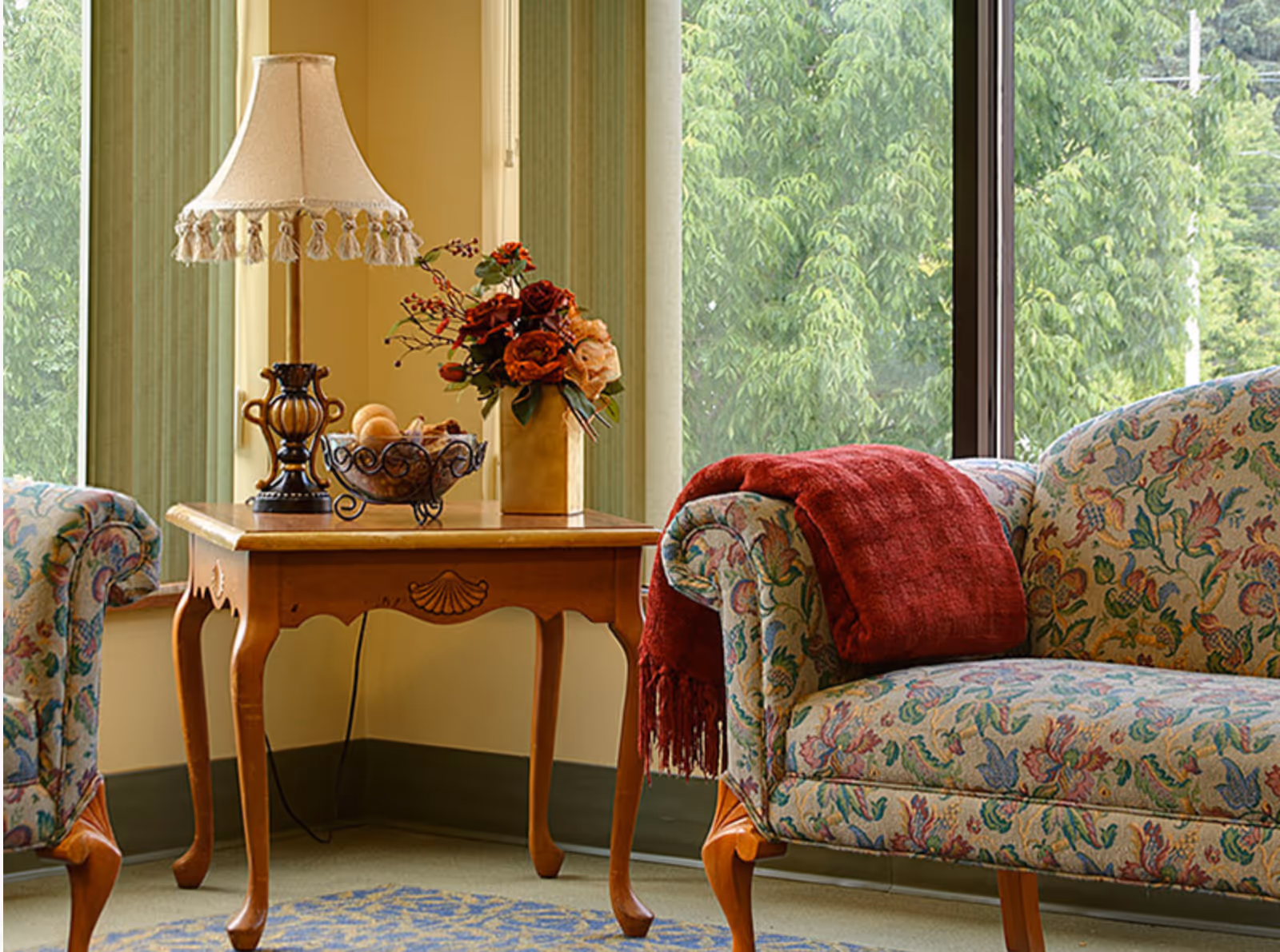 A cozy living room corner with a floral patterned armchair draped with a red throw blanket, a wooden side table holding a decorative lamp, a bowl of fruit, and a vase with flowers, next to large windows showing green trees outside.