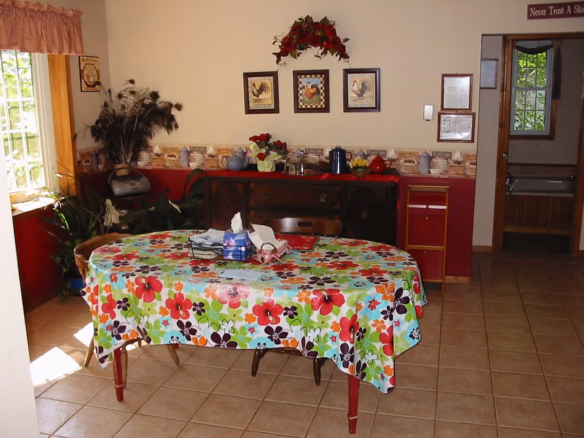 A dining area with a rectangular table covered by a colorful floral tablecloth. On the table are tissue boxes and a few small items. Behind the table is a wooden sideboard with various decorative items, including a vase with flowers and teapots. The walls are decorated with framed pictures of roosters and a floral arrangement above the sideboard. There is a window with a curtain on the left side letting in natural light, and a doorway on the right leading to another room with a bathtub visible.