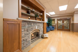 Interior view of a senior living facility hallway with a stone fireplace on the left, a wooden mantle with decorative items, two blue chairs near a large plant, and double glass doors at the end of the hallway.