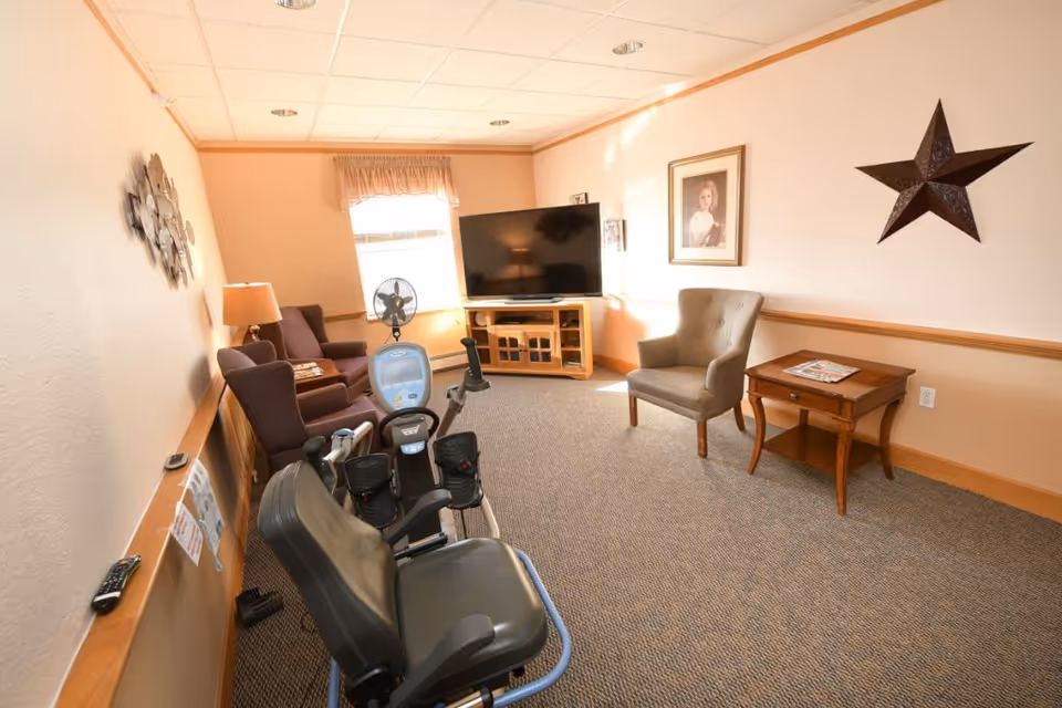 A cozy common living room with armchairs, a TV on a wooden stand, a side table and wall decor, with a recumbent exercise bike in the foreground.