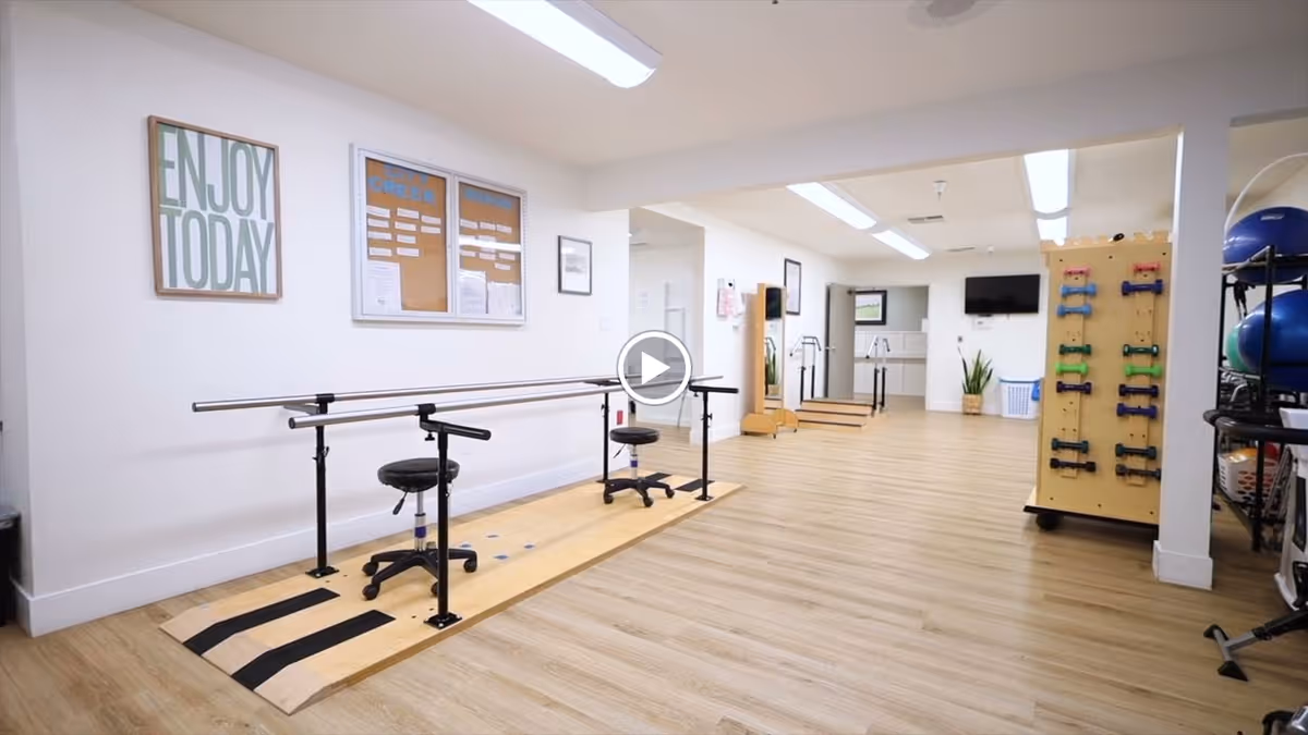 A spacious rehabilitation or physical therapy room with wooden flooring. The room features parallel bars for walking exercises, two black adjustable stools, a rack with colorful dumbbells, exercise balls, and a wall-mounted TV. On the wall, there is a framed sign that reads 'ENJOY TODAY' and a bulletin board with notices. The room is well-lit with ceiling lights and has a clean, organized appearance.
