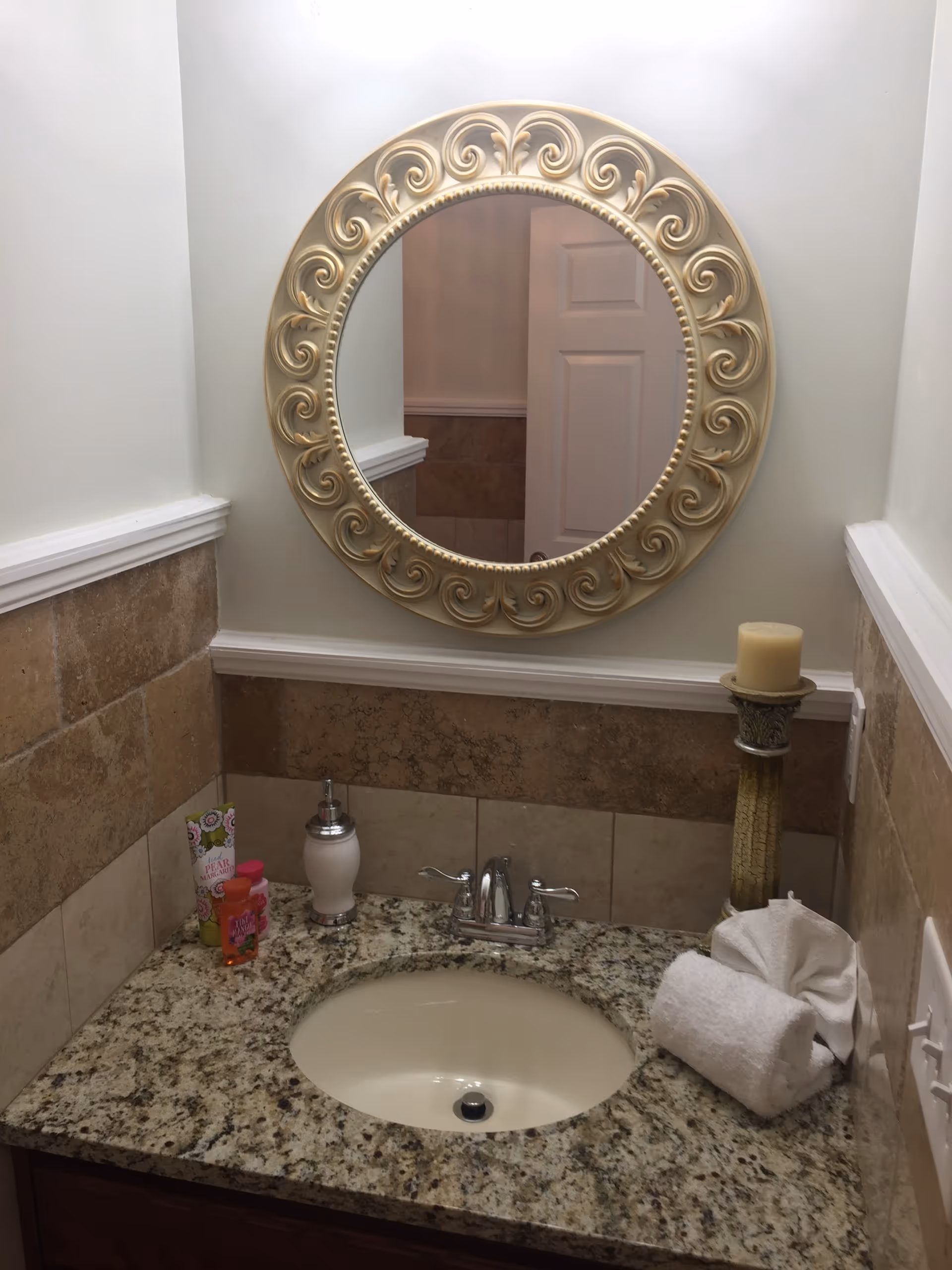 A bathroom sink area with a granite countertop, an oval sink, and a silver faucet. Above the sink is a round decorative mirror with an ornate gold frame. On the countertop, there is a soap dispenser, a small bottle of hand soap, a rolled white towel, a folded white cloth, and a tall candle holder with a beige candle. The walls are tiled halfway up with beige and brown tiles, and the upper half is painted white.