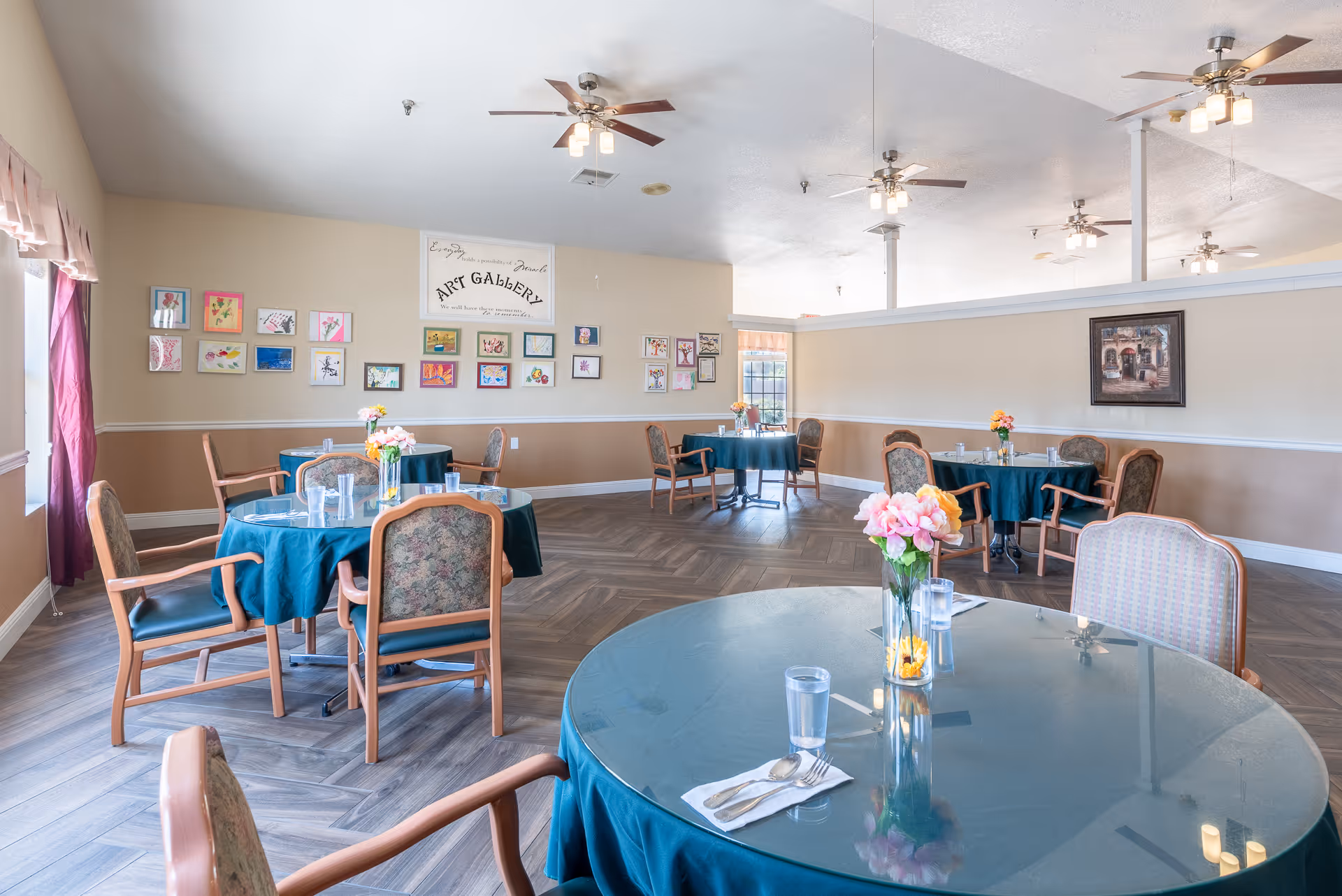 Bright communal dining room with round tables draped in teal tablecloths, floral centerpieces, chairs, and artwork on the walls.