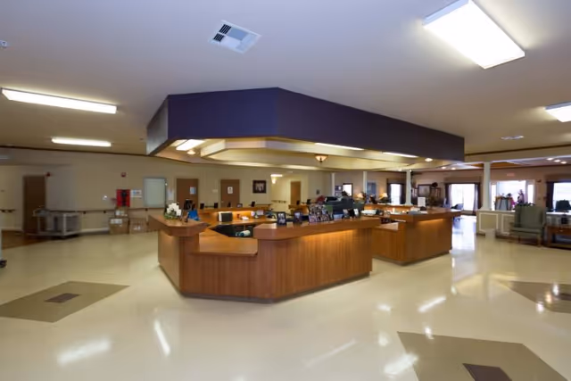 Spacious reception area in a healthcare and rehabilitation facility with a large wooden front desk in the center, surrounded by beige walls and a polished floor. The ceiling has built-in lighting, and there are chairs and windows visible in the background.