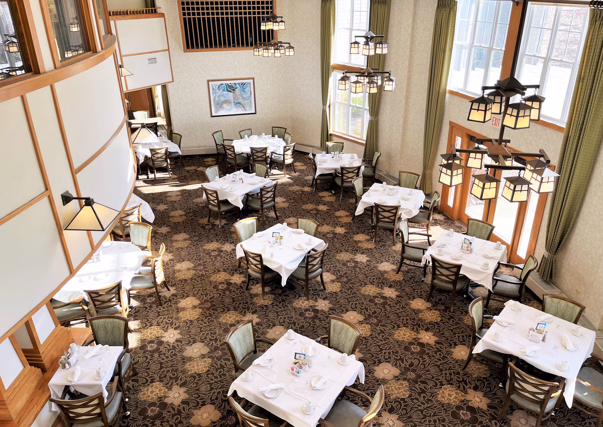 A bright and elegant dining room with multiple tables covered in white tablecloths, set with cups, plates, and napkins. The room features large windows with green curtains, patterned carpet, and hanging lantern-style light fixtures. Chairs with green upholstery surround each table.