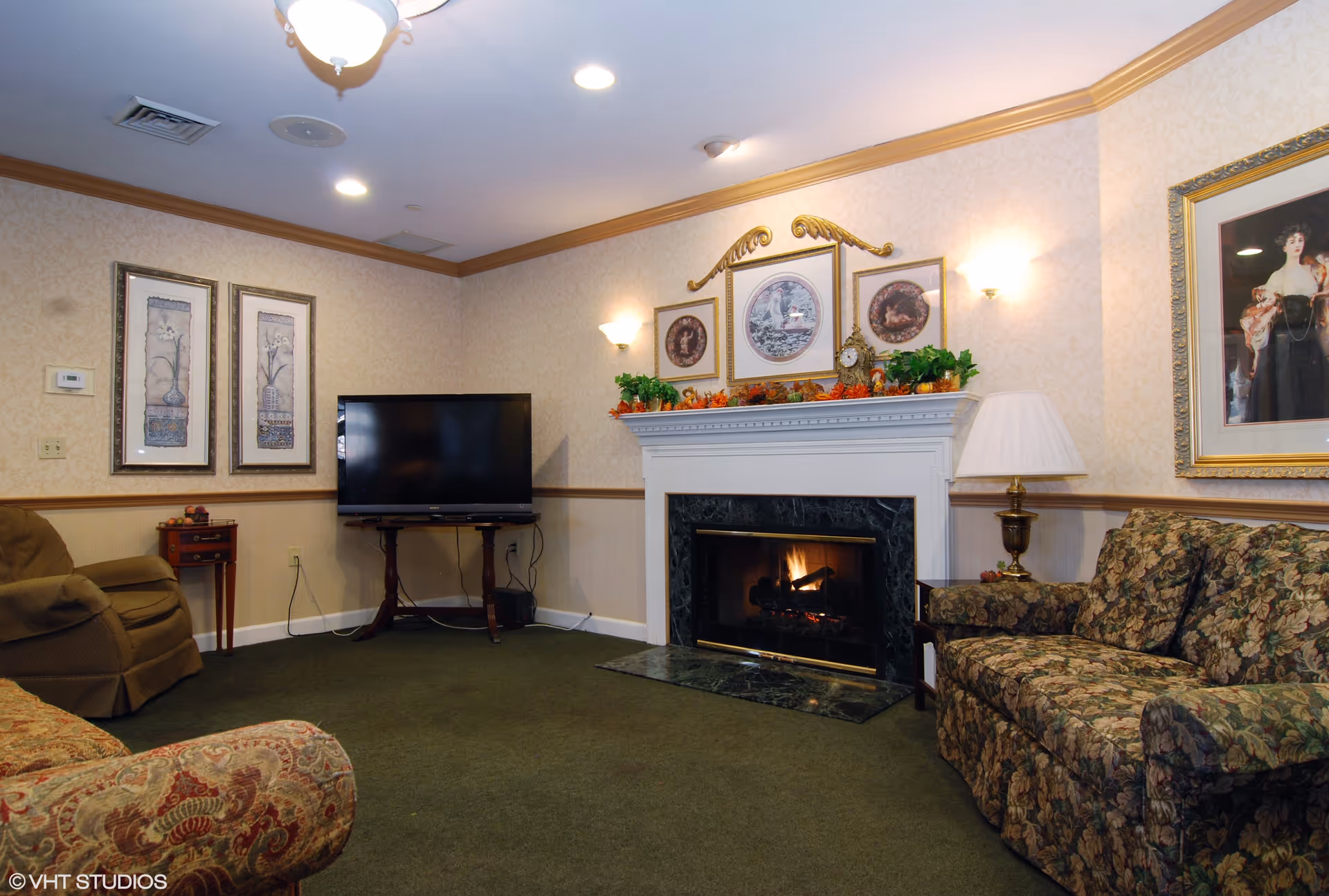 A cozy living room with a green carpet, floral patterned sofa, and armchair. There is a fireplace with a fire burning, decorated with framed pictures and plants on the mantel. A flat-screen TV is placed on a wooden stand in the corner. The walls have a light patterned wallpaper with wooden trim, and there are wall sconces providing warm lighting.