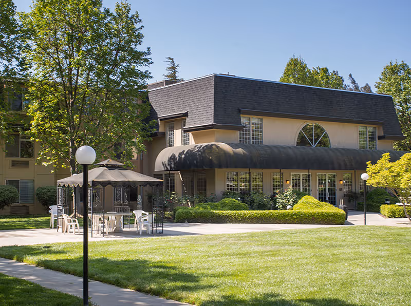 Exterior view of a two-story building with a dark shingled roof and beige walls, surrounded by green trees and shrubs. In front of the building, there is a paved area with a gazebo structure and several white plastic chairs and tables. The scene is set on a sunny day with a well-maintained lawn and a sidewalk leading towards the building.