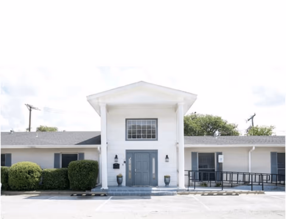 Front entrance of a single-story white building with columns, double doors, shrubs, and a wheelchair ramp.