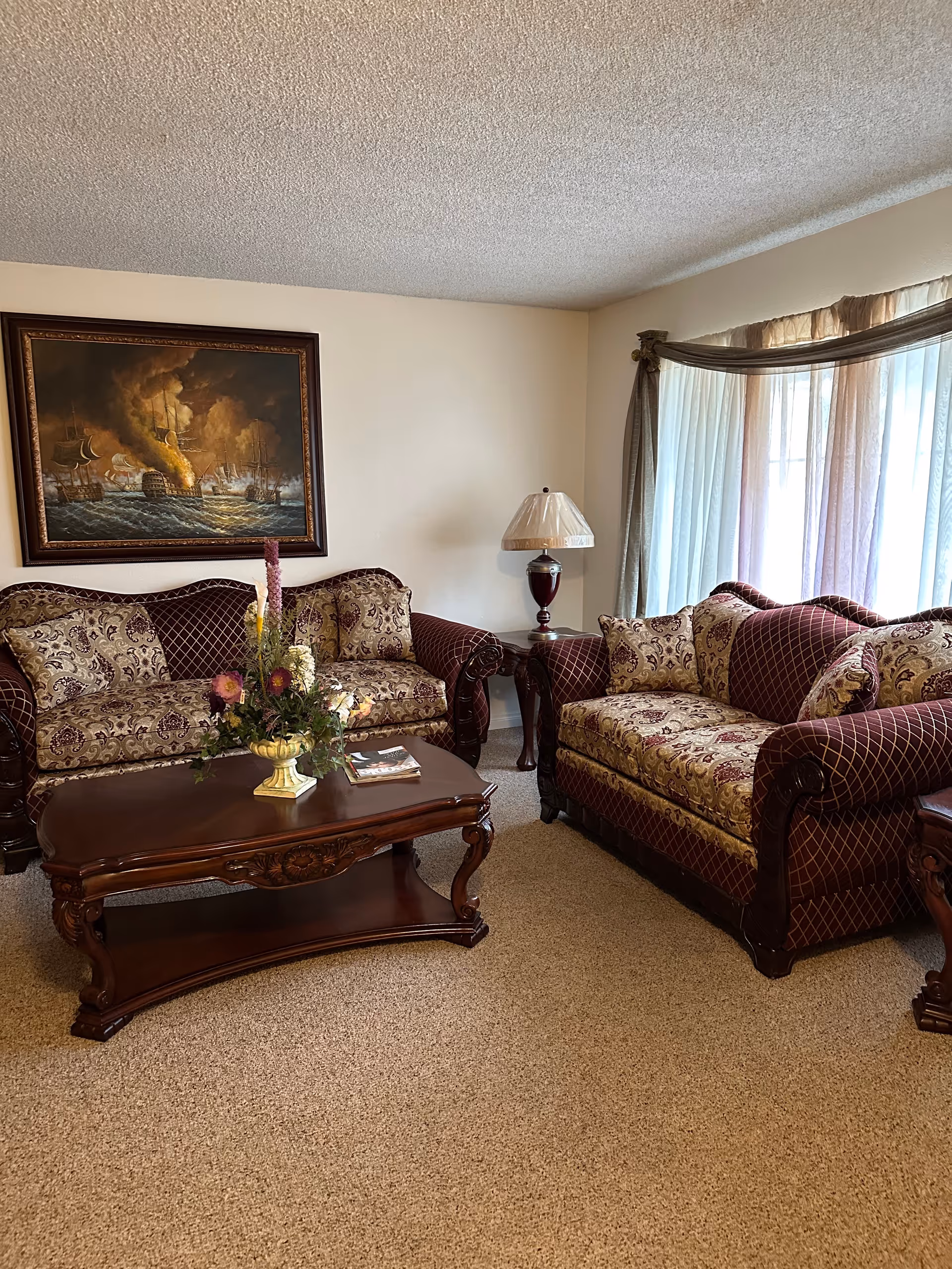 Elegant living room with burgundy patterned sofas, a wood coffee table, lamp, painting, and curtains.
